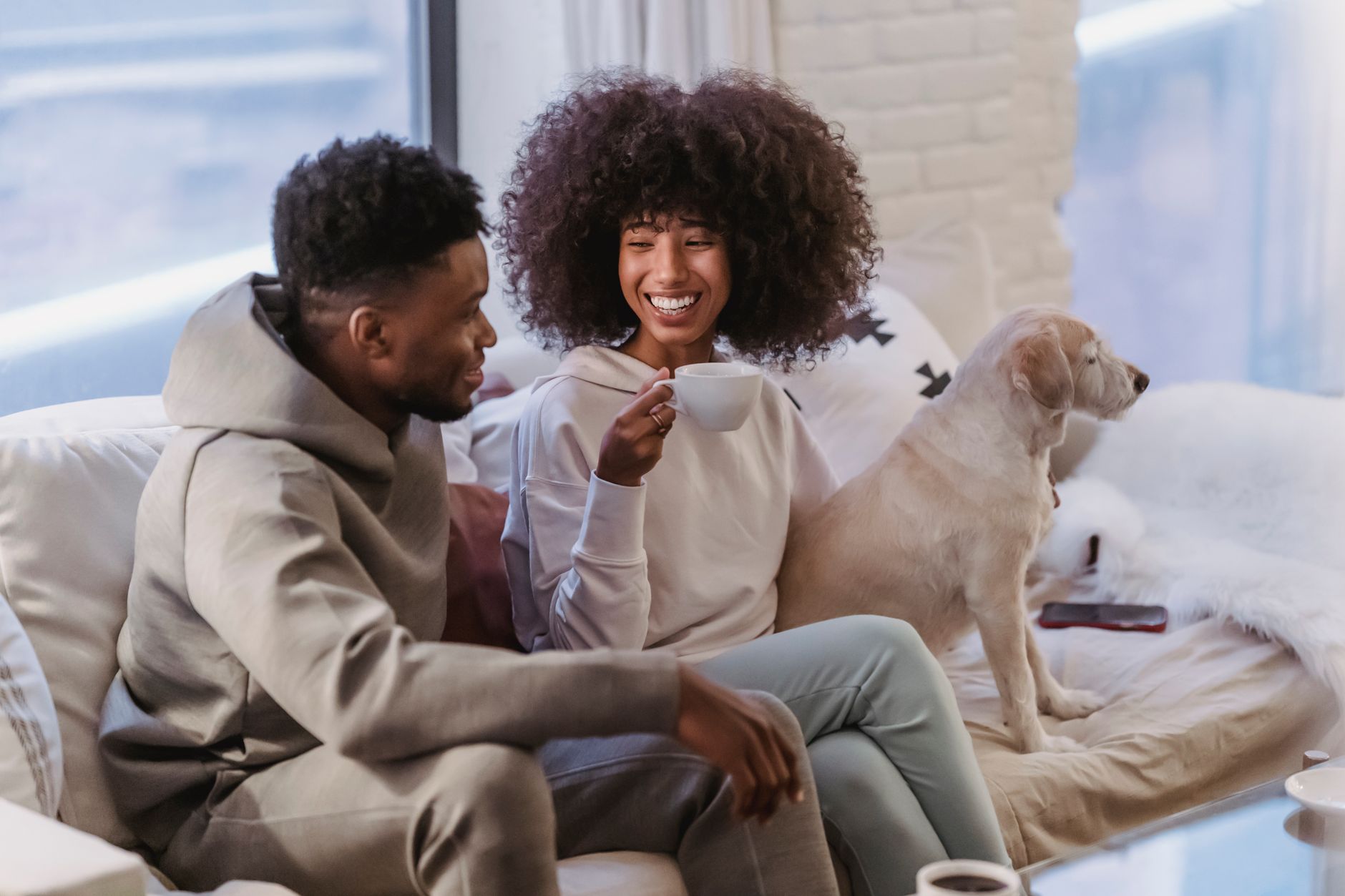 Een stel zit met koffie op de bank en praat lachend met elkaar, met de hond ernaast